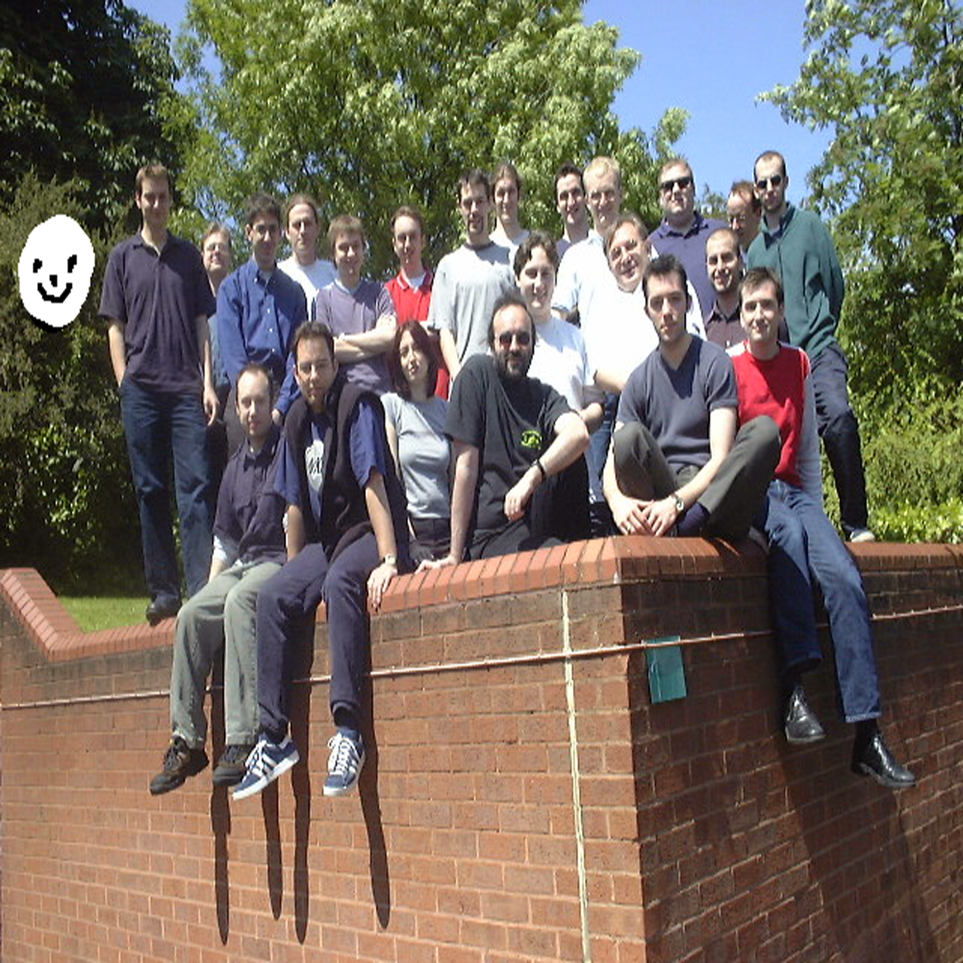A photo of the developers posing on a brick wall with a smiley face crudely drawn to the left.